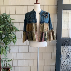 May include: A patchwork jacket with a stand-up collar and ruffled hem. The jacket features a mix of blue, olive green, and burgundy fabrics. Decorative floral patches adorn the shoulders. The jacket is displayed on a mannequin.