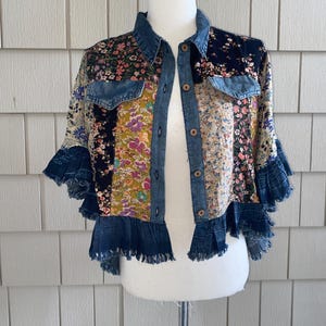 May include: A patchwork jacket with a denim collar, pockets, and trim. The jacket features floral and patterned fabric panels in shades of blue, yellow, pink, and black. The jacket has a button-up front and ruffled sleeves.