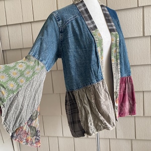 May include: A patchwork jacket featuring a blue denim bodice with long, flared sleeves. The garment incorporates various fabric panels, including green daisy prints, grey plaid, and floral patterns, with a collarless open front design.