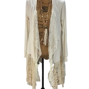 May include: A white cardigan with a lace overlay and a long tassel necklace. The cardigan has a loose fit and a long, flowing design.