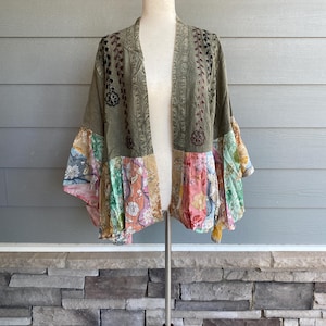 May include: Olive green kimono-style jacket with floral patchwork sleeves. The jacket features embroidered details and a flowing, open front design. The sleeves showcase a variety of colorful floral patterns, adding a bohemian flair to the garment.