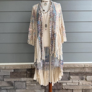 May include: A long, cream-colored lace duster cardigan featuring intricate floral patterns in shades of blue, pink, and brown. The garment has an open front, three-quarter length sleeves, and a high-low hemline with layered lace trim details.