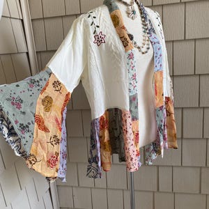 May include: A cream-colored open-front jacket featuring long bell sleeves and a patchwork hem. The garment is accented with embroidered floral details on the shoulders and a multi-colored fabric trim along the front opening and sleeve edges.