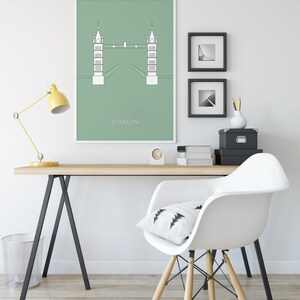 May include: A minimalist green poster with a white outline of the Tower Bridge in London. The poster is framed and hanging on a white wall above a wooden desk with a white chair. The desk has a yellow lamp, a black and white patterned pillow, and a black file box.