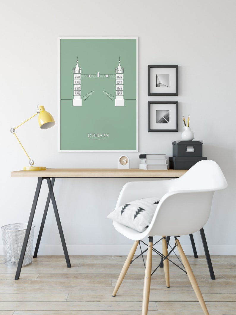 May include: A minimalist green poster with a white outline of the Tower Bridge in London. The poster is framed and hanging on a white wall above a wooden desk with a white chair. The desk has a yellow lamp, a black and white patterned pillow, and a black file box.
