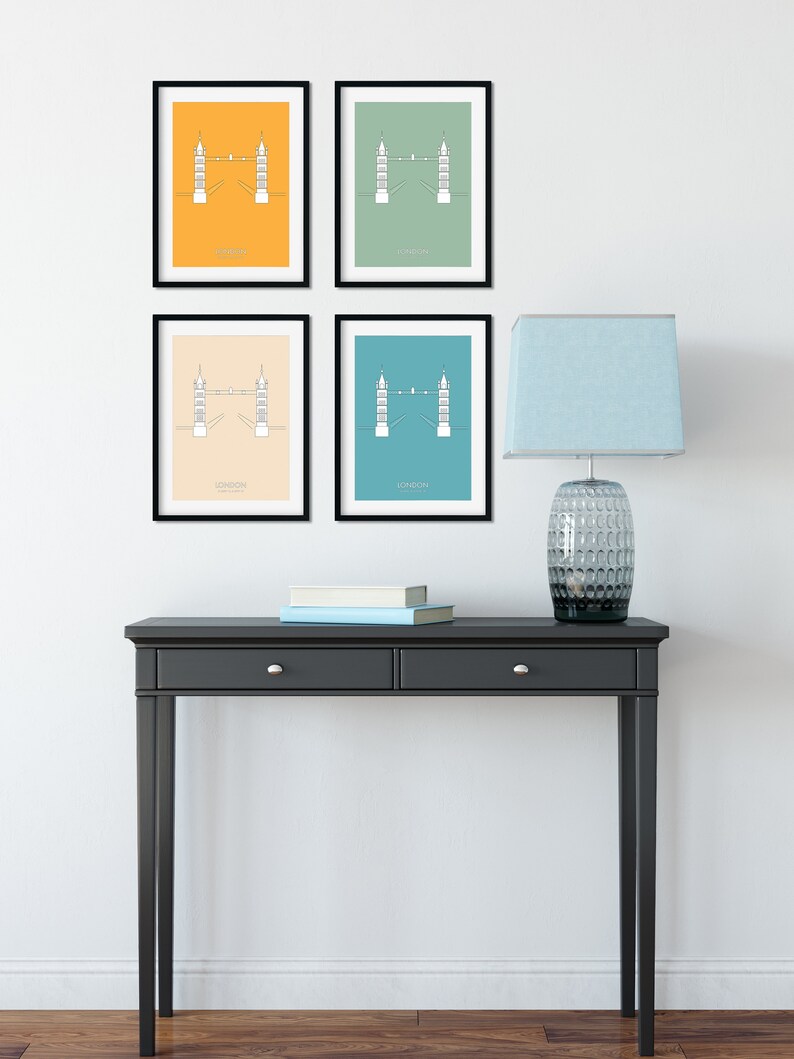 May include: Four framed prints of the Tower Bridge in London, each with a different background color: yellow, green, beige, and blue. The prints are hanging on a white wall above a black console table with two drawers. A blue lamp with a clear glass base is on the table.