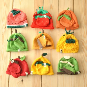 May include: A collection of knitted hats in various fruit designs, including a watermelon, strawberry, carrot, and pineapple. Each hat has a matching pocket and is displayed against a light wood background. The hats are colorful and whimsical.