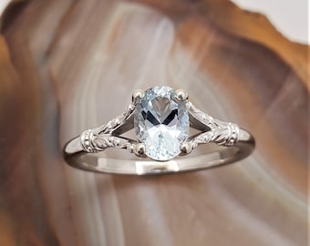 Puede incluir: Un anillo de plata con una piedra preciosa ovalada de color azul claro. El anillo tiene un diseño calado con detalles florales en la banda. La piedra preciosa está engastada con cuatro garras. El anillo se muestra sobre un fondo neutro.