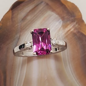 May include: A silver ring featuring a large, rectangular, pink gemstone and two smaller, round, white gemstones.