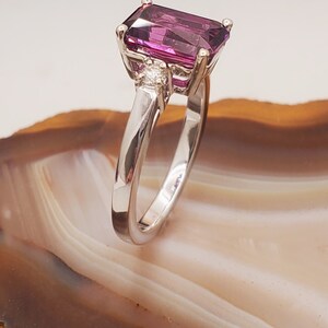 May include: A silver ring featuring a large, rectangular, purple gemstone and two smaller, round, white gemstones.