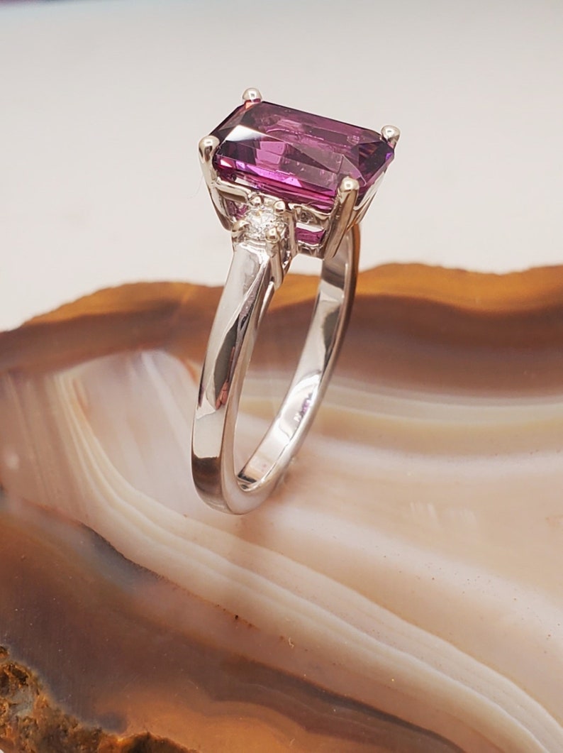 May include: A silver ring featuring a large, rectangular, purple gemstone and two smaller, round, white gemstones.