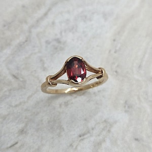 May include: A gold ring featuring a vibrant oval-cut red gemstone. The ring's band has a delicate, openwork design that cradles the gemstone. The ring is set against a neutral background, highlighting its elegant simplicity.