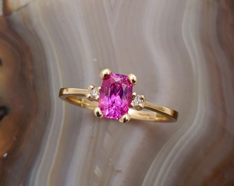 May include: A gold ring with a pink gemstone and two smaller diamonds. The ring is set on a gray and white background.