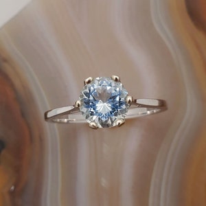 May include: A solitaire engagement ring featuring a large, round, light blue gemstone. The gemstone is set in a gold-colored six-prong setting, with a thin, silver-colored band. The ring is displayed against a neutral, swirled background.