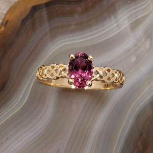 Puede incluir: Un anillo de oro con una piedra ovalada rosa engastada en un dise&ntilde;o de nudo celta.