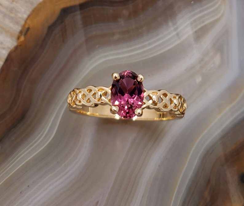 Puede incluir: Un anillo de oro con una piedra ovalada rosa engastada en un dise&ntilde;o de nudo celta.