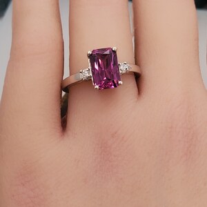 May include: A ring featuring a large, rectangular, dark purple gemstone surrounded by smaller, clear gemstones. The ring is set in white metal.