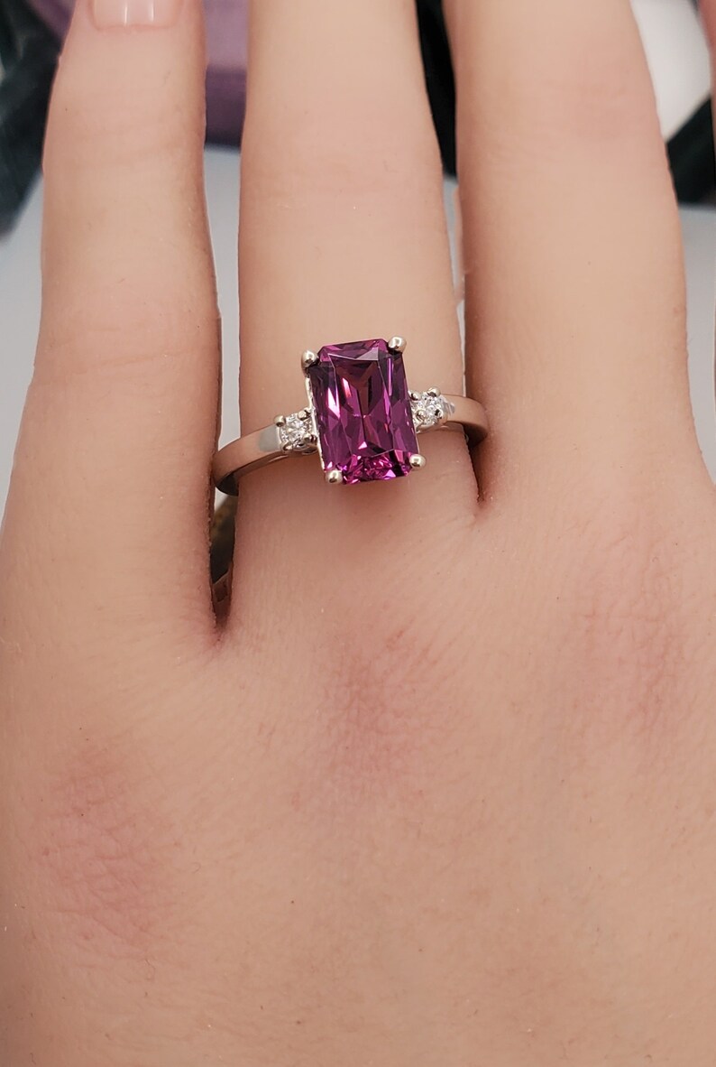May include: A ring featuring a large, rectangular, dark purple gemstone surrounded by smaller, clear gemstones. The ring is set in white metal.
