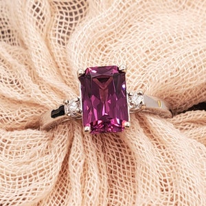 May include: A silver ring featuring a large, rectangular, purple gemstone and two smaller, round, white gemstones set into the band.
