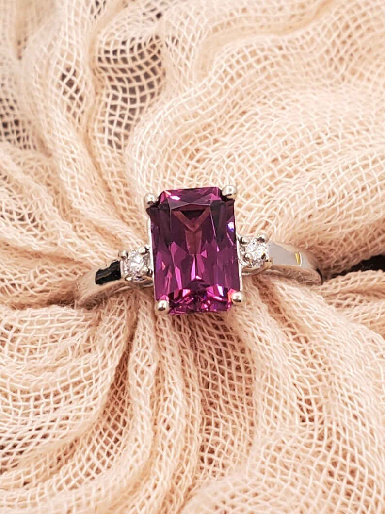 May include: A silver ring featuring a large, rectangular, purple gemstone and two smaller, round, white gemstones set into the band.