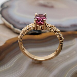 Puede incluir: Un anillo de oro con una piedra preciosa rosa engastada en un dise&ntilde;o delicado y ornamentado. El anillo est&aacute; sobre un fondo blanco y marr&oacute;n.