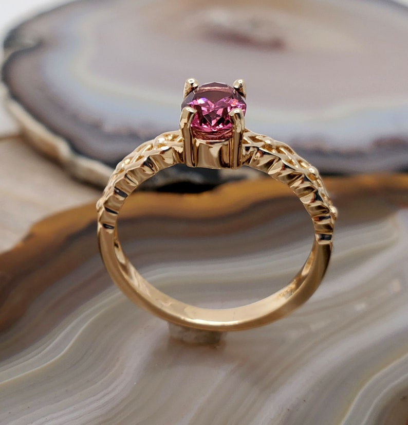 Puede incluir: Un anillo de oro con una piedra preciosa rosa engastada en un dise&ntilde;o delicado y ornamentado. El anillo est&aacute; sobre un fondo blanco y marr&oacute;n.