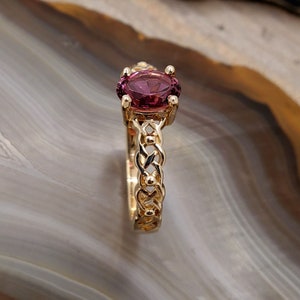 Puede incluir: Un anillo de oro con una banda trenzada y una piedra ovalada de color p&uacute;rpura.