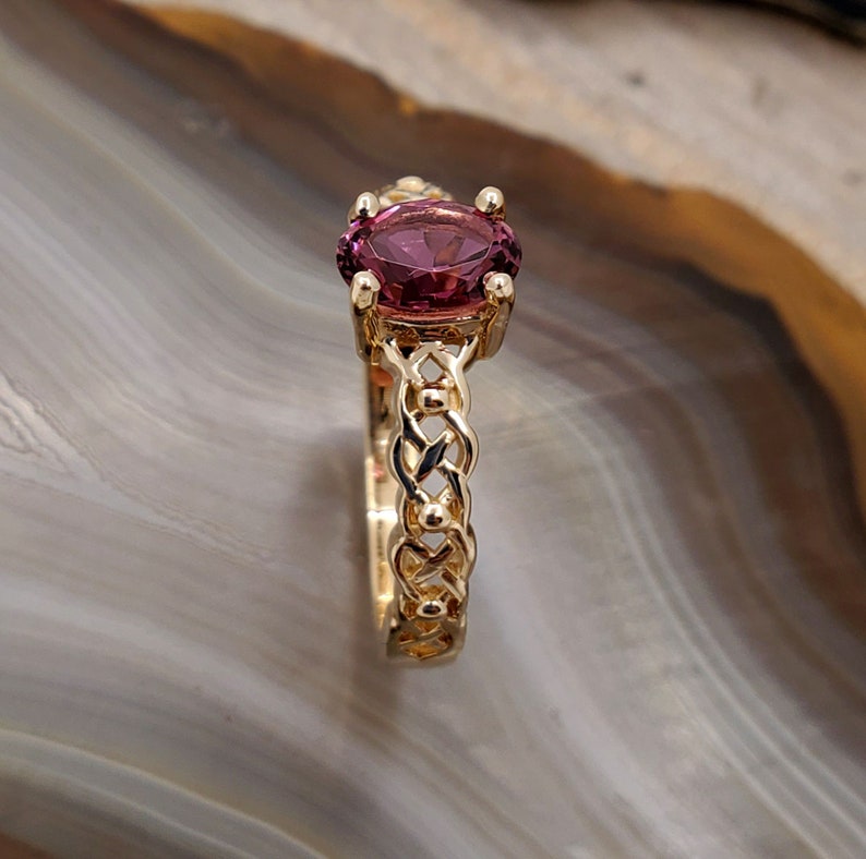Puede incluir: Un anillo de oro con una banda trenzada y una piedra ovalada de color p&uacute;rpura.