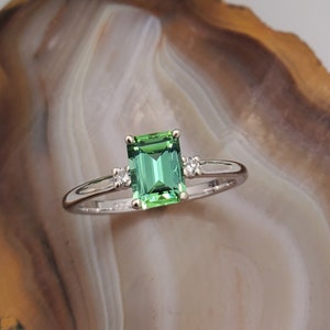 May include: A silver ring with a green emerald-cut gemstone and two smaller diamonds on the band.