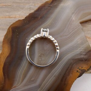 May include: A silver ring with a single clear stone set in the center. The band is decorated with small, evenly spaced stones.