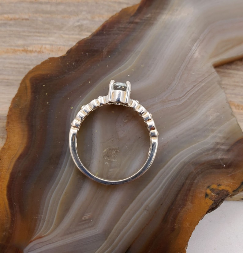 May include: A silver ring with a single clear stone set in the center. The band is decorated with small, evenly spaced stones.