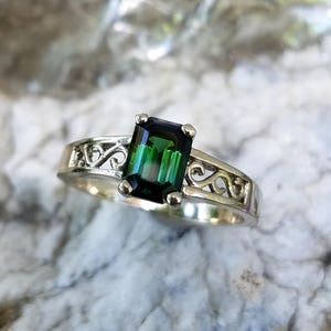 May include: A silver ring with a green emerald-cut gemstone set in a delicate filigree design.