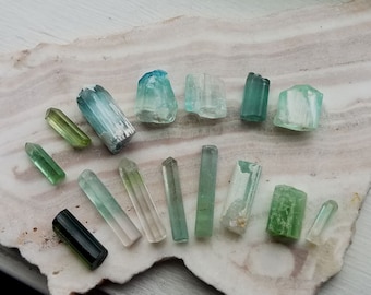 May include: A collection of 14 green and blue aquamarine crystals of varying sizes and shapes. The crystals are arranged on a white and green surface.