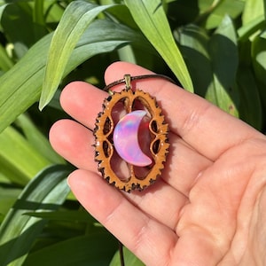 May include: A pendant featuring a crescent moon shape with an iridescent, pearlescent finish set inside a natural, textured brown walnut shell casing. The piece is attached to a dark brown cord, creating a necklace with a distinct organic aesthetic.