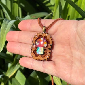 May include: A pendant necklace featuring a small, iridescent mushroom figurine set inside a brown, textured walnut shell half. The pendant is attached to a brown leather cord, creating a natural, earthy accessory with a glossy, colorful finish.