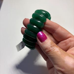 May include: A stretch bracelet composed of multiple smooth, rounded, dark green translucent beads. The beads are strung together in a continuous circular loop, creating a uniform, polished appearance with a small white rectangular tag attached.