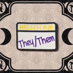 May include: A rectangular embroidered patch featuring a white background with a black border. The text HELLO I AM is stitched in yellow at the top, and the text They/Them is stitched in purple across the center of the patch.