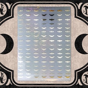May include: A sheet of silver and gold bat-shaped stickers. The stickers are arranged in rows and columns on a light blue background with a subtle glitter effect. The sheet is framed by a decorative border with crescent moons and the letter "N".