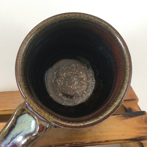 以下が含まれることがあります： 手作りのセラミックマグカップ。内側は濃い茶色、外側は赤みがかった茶色。ハンドルは青、緑、紫の色合いが混ざっています。マグカップは木製の表面に置かれています。