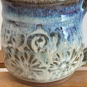 Pu&ograve; includere: Una tazza in ceramica fatta a mano con una smaltatura unica. La tazza presenta una miscela di tonalit&agrave; blu, bianche e marroni, con un design strutturato e modellato all'esterno. Ha un manico e una base arrotondata, perfetta per bevande calde.