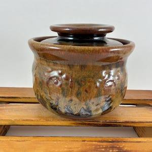 May include: A round ceramic butter crock with a lid, featuring a glossy glaze in shades of brown, tan, and dark amber. The exterior surface has a mottled, textured appearance with subtle raised circular patterns around the base.