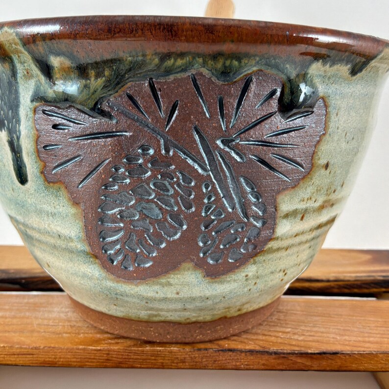 May include: A ceramic bowl with a rounded shape, featuring a glazed finish in shades of brown and light green. The exterior displays a dark brown, embossed design of two pinecones and pine needles centered on the side of the bowl.