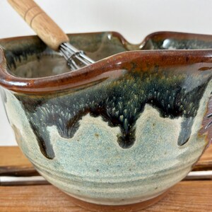 May include: A ceramic mixing bowl with a scalloped rim and a reactive glaze featuring a light blue base with dark brown and green drips cascading from the top edge. A metal whisk with a light-colored wooden handle rests inside the bowl.