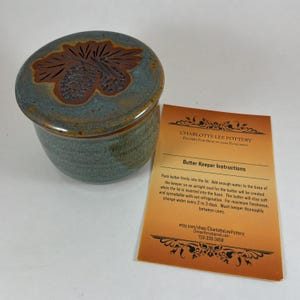 May include: A round ceramic butter keeper with a blue-green glaze and a lid featuring a carved pinecone design. Beside it is an orange instruction card titled Charlotte Lee Pottery Butter Keeper Instructions with text detailing usage and care.