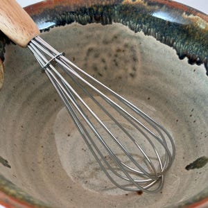 May include: A stainless steel wire whisk with a light-colored wooden handle resting inside a speckled ceramic bowl. The bowl features a cream-colored interior and a dark, textured rim with brown and green tones along the outer edge.