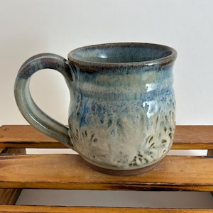Pu&ograve; includere: Tazza in ceramica fatta a mano con corpo arrotondato e grande manico. La tazza presenta una glassa blu e bianca con una superficie strutturata. Il bordo e il manico sono di una tonalit&agrave; di blu pi&ugrave; scura. La tazza &egrave; appoggiata su una superficie di legno.