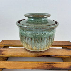 May include: A ceramic butter crock with a lid, featuring a glossy glaze that transitions from a sage green at the top to a warm, earthy brown at the base. The exterior surface has a textured, vertical drip pattern throughout the body.
