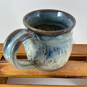 Pu&ograve; includere: Tazza in ceramica fatta a mano con un design marmorizzato unico nei toni del blu, bianco e marrone. La tazza presenta un manico grande e curvo e un bordo leggermente svasato. L'interno e il manico hanno una finitura lucida.