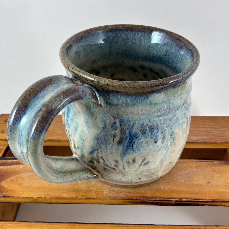 Pu&ograve; includere: Tazza in ceramica fatta a mano con un design marmorizzato unico nei toni del blu, bianco e marrone. La tazza presenta un manico grande e curvo e un bordo leggermente svasato. L'interno e il manico hanno una finitura lucida.