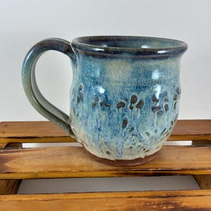 May include: A handmade ceramic mug with a blue and brown glaze. The mug has a rounded shape with a handle and a textured surface. The interior is a lighter blue, and the rim is a darker shade of blue.